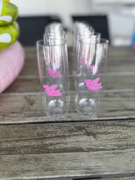 Row of clear plastic champagne flutes with pink script logo lined up on a weathered wooden outdoor table — shallow depth-of-field party glassware