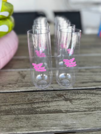 Row of clear plastic champagne flutes with pink script logo lined up on a weathered wooden outdoor table — shallow depth-of-field party glassware