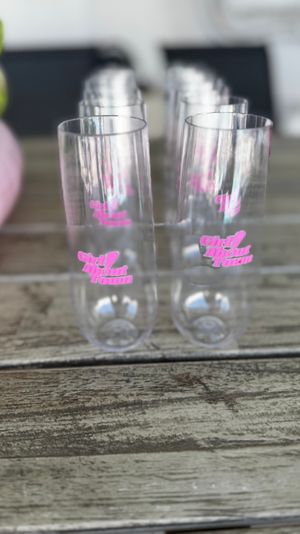 Row of clear plastic champagne flutes with pink script logo lined up on a weathered wooden outdoor table — shallow depth-of-field party glassware