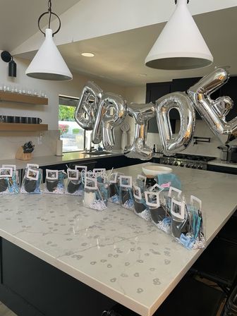 Modern kitchen island with marble countertop decorated for a bridal shower — large silver 'BRIDE' foil balloons and a row of clear party favor bags lined up under white pendant lights