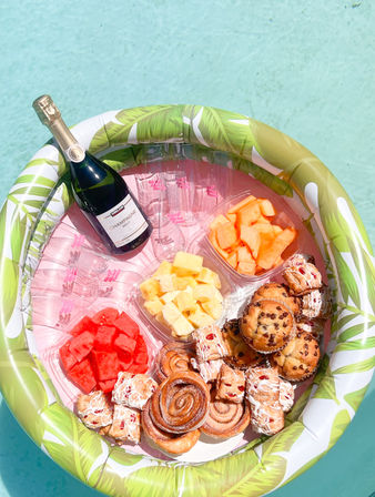 Tropical inflatable pool float in turquoise water holding a bottle of champagne, plastic cups and a colorful poolside brunch spread with watermelon, pineapple, cantaloupe, cinnamon rolls, muffins and glazed pastries.