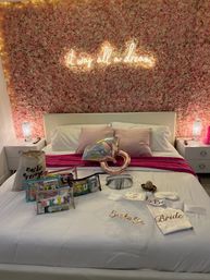 Pink floral feature wall with glowing neon sign above a white bed staged for a bridal celebration, satin 'Bride' robe, party favors, and rose-gold heart balloon on crisp white linens