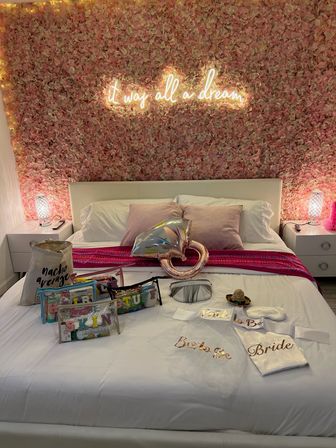 Pink floral feature wall with glowing neon sign above a white bed staged for a bridal celebration, satin 'Bride' robe, party favors, and rose-gold heart balloon on crisp white linens