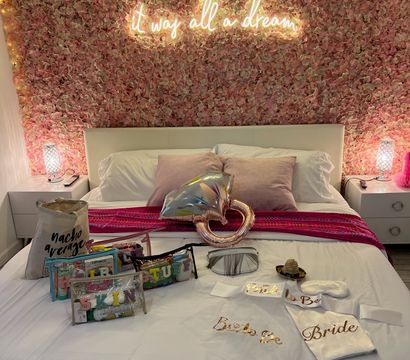 Pink floral feature wall with glowing neon sign above a white bed staged for a bridal celebration, satin 'Bride' robe, party favors, and rose-gold heart balloon on crisp white linens