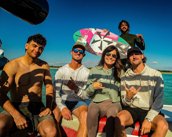 Five people on a boat with a colorful wakesurf board, smiling and flashing shaka signs under a bright blue sky over turquoise coastal water.