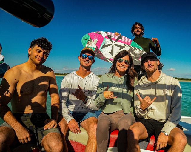 Five people on a boat with a colorful wakesurf board, smiling and flashing shaka signs under a bright blue sky over turquoise coastal water.