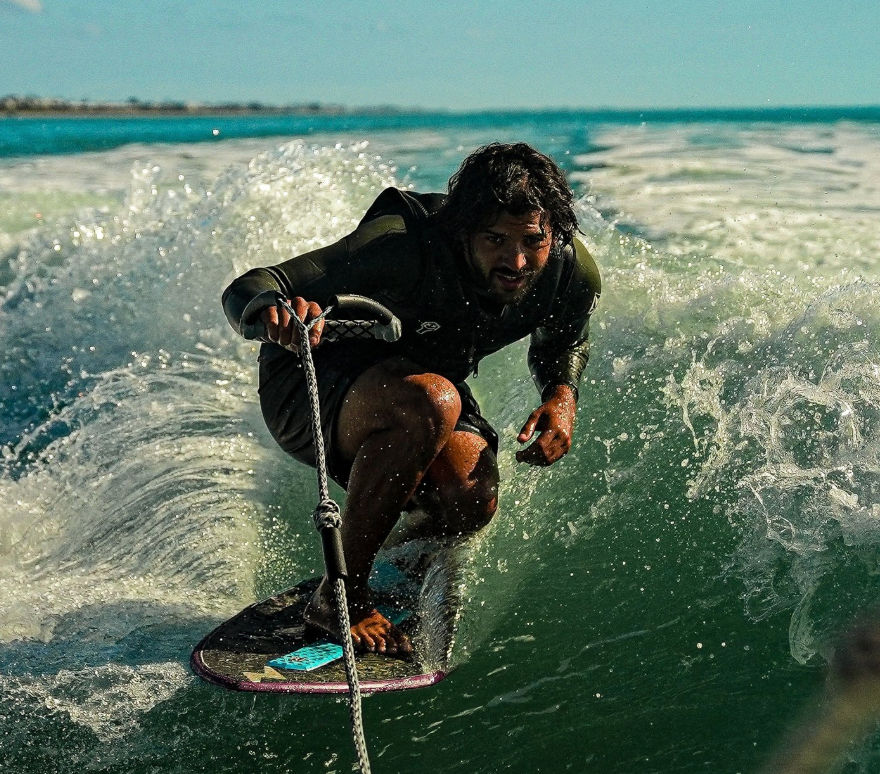 Wakesurfer crouched low on a board, gripping a tow rope and carving through a sunlit boat wake in turquoise coastal ocean waters — high-energy water sports action.