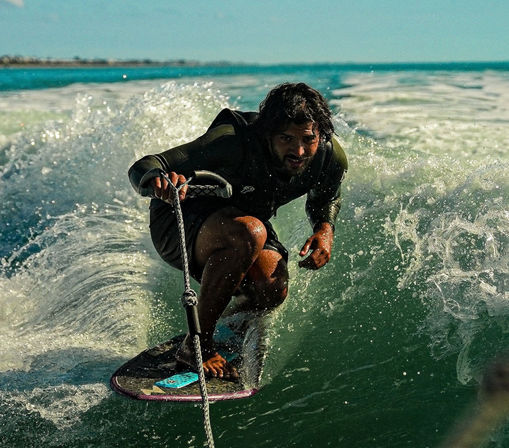 Wakesurfer crouched low on a board, gripping a tow rope and carving through a sunlit boat wake in turquoise coastal ocean waters — high-energy water sports action.