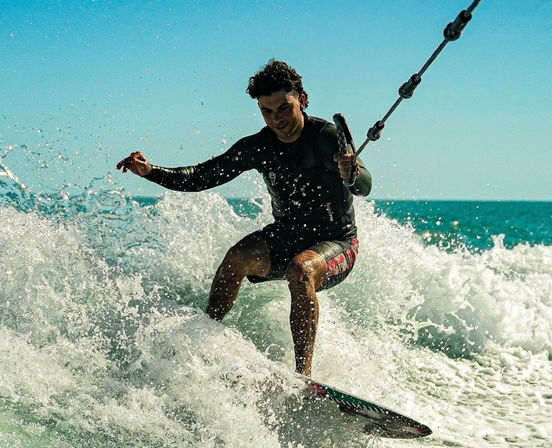Wakeboarder gripping a tow rope and carving through white surf on turquoise ocean under a clear sunny sky