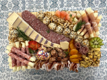 Overhead shot of a colorful charcuterie board on a patterned tablecloth: sliced cheeses and salami, prosciutto-wrapped cheese sticks, sesame-coated cheese balls, dried apricots, strawberries, kiwi, walnuts and candied nuts — vibrant cheese and meat grazing board for parties.