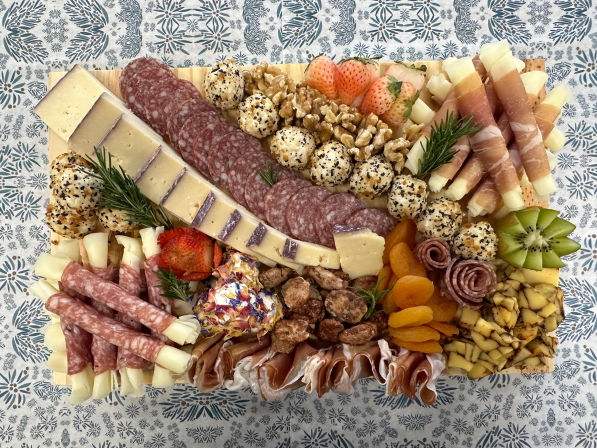 Overhead shot of a colorful charcuterie board on a patterned tablecloth: sliced cheeses and salami, prosciutto-wrapped cheese sticks, sesame-coated cheese balls, dried apricots, strawberries, kiwi, walnuts and candied nuts — vibrant cheese and meat grazing board for parties.