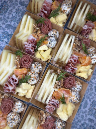 Overhead shot of artisan charcuterie boxes arranged for catering — sliced cheeses, rolled prosciutto and salami roses, seed-crusted cheese balls, dried apricots, walnuts and rosemary sprigs on a patterned tablecloth, perfect for picnics or events.