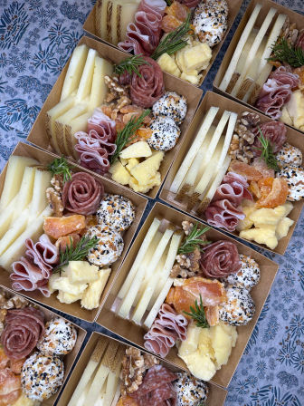 Overhead shot of artisan charcuterie boxes arranged for catering — sliced cheeses, rolled prosciutto and salami roses, seed-crusted cheese balls, dried apricots, walnuts and rosemary sprigs on a patterned tablecloth, perfect for picnics or events.