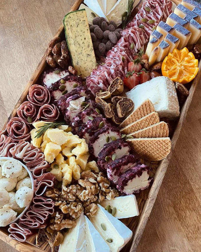 Gourmet wooden charcuterie board with assorted cheeses (brie, goat logs, hard cheddar), rolled salami and prosciutto, wafer crackers, figs, walnuts, strawberries, mandarin slices, marinated mozzarella, and seed-topped cheese rounds.