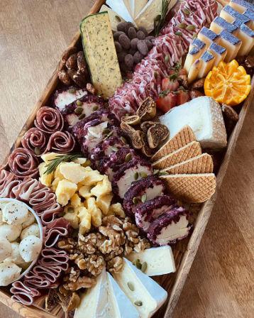 Gourmet wooden charcuterie board with assorted cheeses (brie, goat logs, hard cheddar), rolled salami and prosciutto, wafer crackers, figs, walnuts, strawberries, mandarin slices, marinated mozzarella, and seed-topped cheese rounds.
