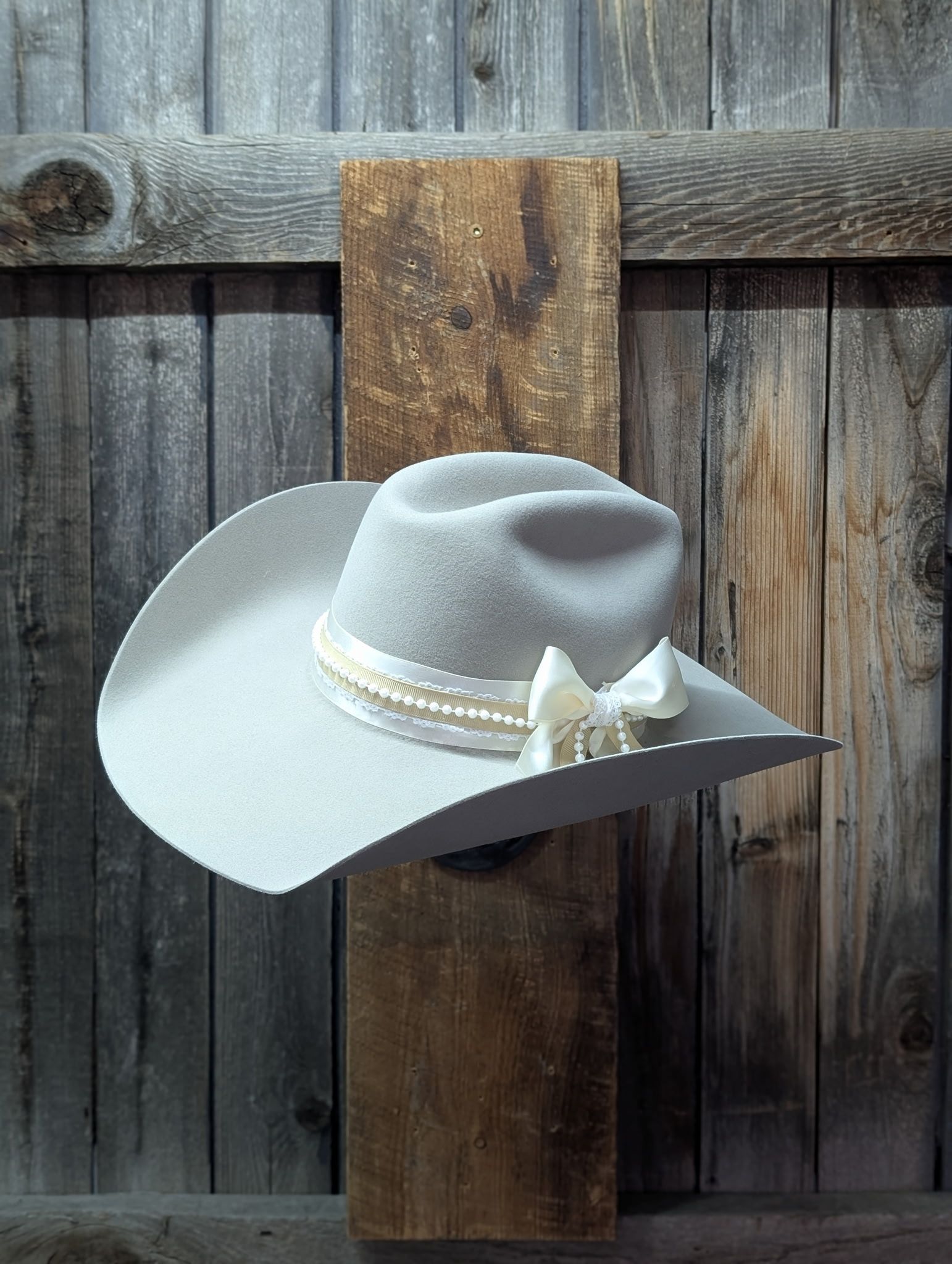 Cream felt cowboy hat with ivory ribbon, pearl trim and bow displayed against a rustic barn-wood backdrop