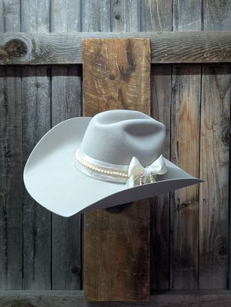 Cream felt cowboy hat with ivory ribbon, pearl trim and bow displayed against a rustic barn-wood backdrop