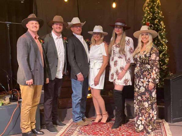 Six adults wearing cowboy hats and semi-formal country‑western outfits smile while posing on a small indoor stage with string lights, a decorated Christmas tree, and a red patterned rug.