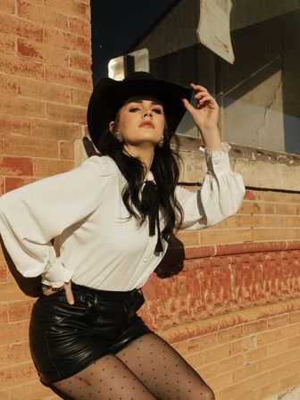 Sunlit urban fashion portrait of a person in a black cowboy hat, white blouse with a black bow, leather shorts and polka-dot tights leaning against a red brick building — downtown cowgirl-inspired street style.