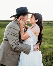 Romantic outdoor wedding portrait: bride in an off-shoulder lace gown and groom in a suit with a black cowboy hat embrace, smiling nose-to-nose in a sunlit grassy country field at sunset.