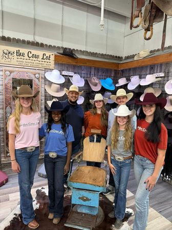 Smiling group wearing cowboy hats pose inside a western-style hat shop with a colorful hat wall, vintage barber chair, and cowhide rug.