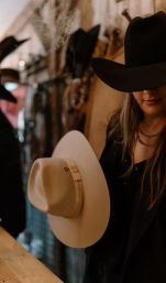 Person wearing a black cowboy hat holding a beige wide-brimmed western hat in a rustic hat shop, warm moody lighting and country vibe.
