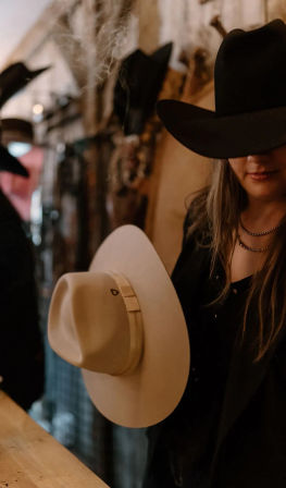 Person wearing a black cowboy hat holding a beige wide-brimmed western hat in a rustic hat shop, warm moody lighting and country vibe.