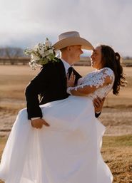 Newlyweds in a sunlit outdoor wedding portrait — groom in a suit and cowboy hat lifts a smiling bride in a lace gown holding a white bouquet in a golden ranch field
