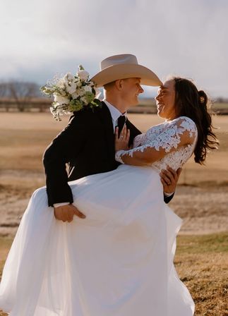 Newlyweds in a sunlit outdoor wedding portrait — groom in a suit and cowboy hat lifts a smiling bride in a lace gown holding a white bouquet in a golden ranch field
