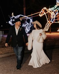 Couple in cowboy hats — bride in a white gown and fur wrap and groom in a blazer and jeans — walking hand‑in‑hand at night past illuminated wire‑frame western light sculptures of bulls and horses.
