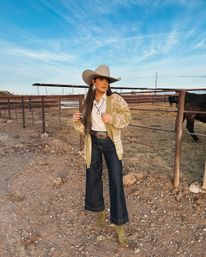 Stylish cowgirl in a beige cowboy hat, patterned cardigan, white shirt, wide-leg denim and green cowboy boots posing by a metal cattle corral on a ranch under a blue sky
