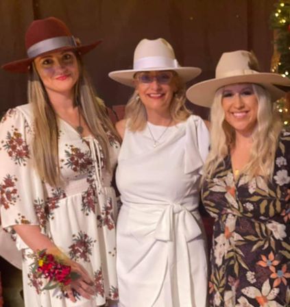 Three smiling women in wide-brim hats posing indoors at a boho country-chic celebration; two wear floral dresses and the center woman wears a white belted dress, one holds a small bouquet.