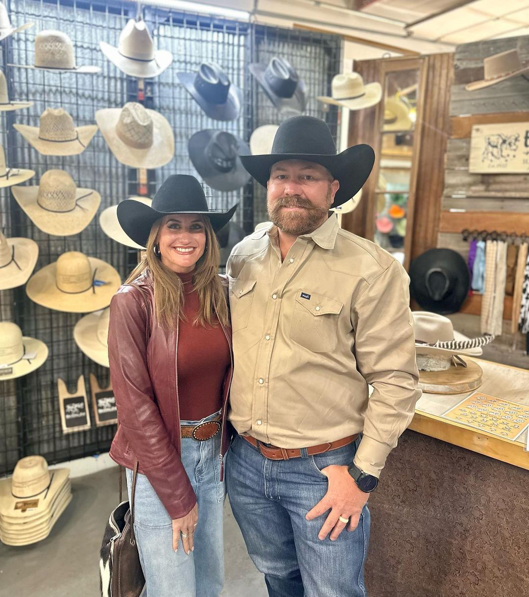 Smiling man and woman wearing black cowboy hats pose inside a western hat shop with rows of straw and felt hats on display.