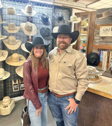 Smiling man and woman wearing black cowboy hats pose inside a western hat shop with rows of straw and felt hats on display.