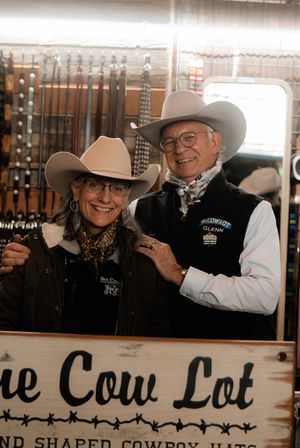 Two smiling people in cowboy hats and western wear pose inside a rustic tack shop with leather bridles on the wall and a wooden sign in the foreground.