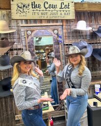 Two smiling pageant contestants in cowboy hats and sashes pose in a rustic western hat shop in front of a decorative mirror, surrounded by displayed cowboy hats and wooden decor.