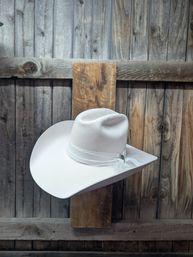 White felt cowboy hat with decorative lace band hanging on a rustic wooden plank fence against a weathered barn wall