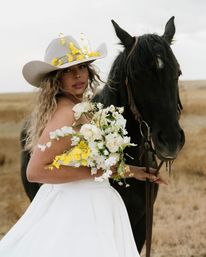 Cowgirl bride in a white dress and flower-adorned cowboy hat holding a white and yellow bridal bouquet beside a black horse in a rustic prairie field, western wedding portrait
