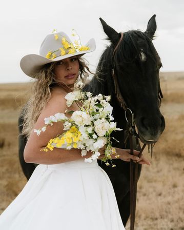 Cowgirl bride in a white dress and flower-adorned cowboy hat holding a white and yellow bridal bouquet beside a black horse in a rustic prairie field, western wedding portrait