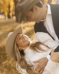 Romantic autumn portrait of a smiling couple in wide-brimmed cowboy hats, woman in white dress and gold earrings and man in a suit embracing in a golden fall field