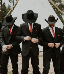 Three groomsmen in black suits and cowboy hats with red ties and pocket squares buttoning jackets in front of a white teepee at an outdoor woodland western wedding.