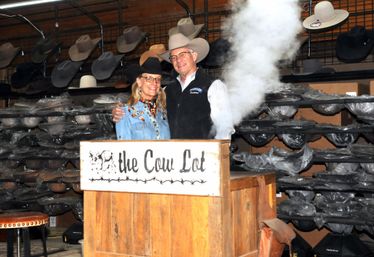Smiling man and woman in cowboy hats stand behind a wooden counter inside a western hat shop with stacked hat racks and a plume of steam rising from a display.
