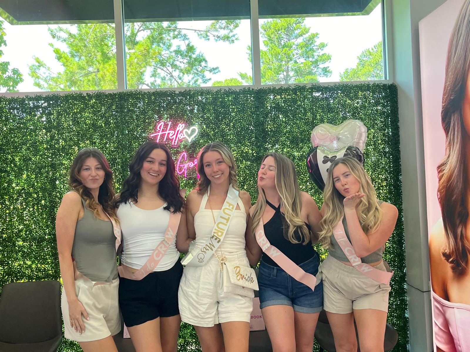 Five women posing at an indoor bridal shower/bachelorette party in front of a faux greenery wall and pink neon sign; bride-to-be in center wearing white with a 'Future Mrs' sash and 'Bride' bag, friends in casual summer outfits and pink sashes, one blowing a kiss, heart-shaped balloons overhead.
