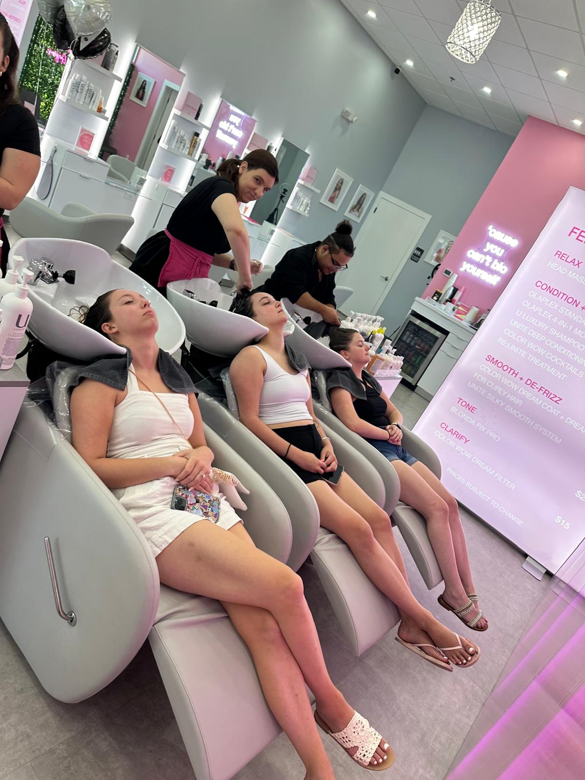 Three women reclining in salon shampoo chairs getting hair washed by stylists in a chic pink-and-gray boutique salon with product shelves and a neon service menu.