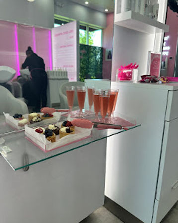 Modern pink-accented beauty salon interior with glass counter displaying mini desserts and flutes of rosé, and a stylist at a hair‑wash station in the background.