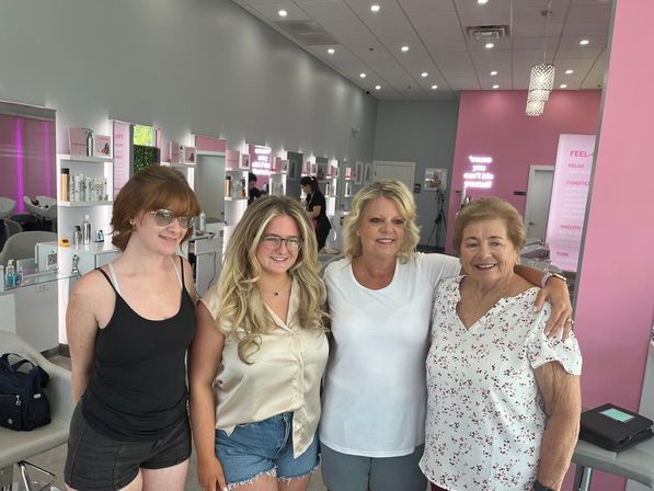 Four smiling women of different ages posing inside a bright modern hair salon with pink accents, styling stations, mirrors and shampoo chairs