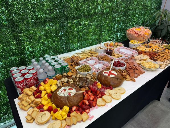 Catered charcuterie buffet spread with breads, crackers, cheeses, cured meats, olives, dips, fresh fruit, shrimp cocktail and canned sodas on a table against a green hedge backdrop.