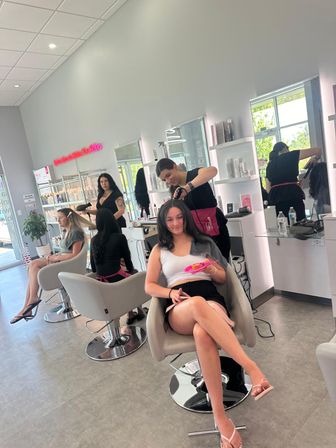 Smiling client lounging in a modern bright hair salon while a stylist straightens her hair; row of gray styling chairs, mirrors, product shelves, large windows and a neon accent sign.