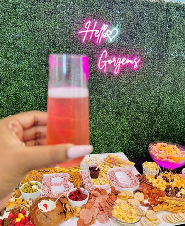 Hand holding a pink cocktail flute in front of a lush green hedge wall with a neon “Hello Gorgeous” sign above a colorful charcuterie grazing table of cheeses, crackers, fruit, macarons and shrimp—cheerful garden brunch vibe.