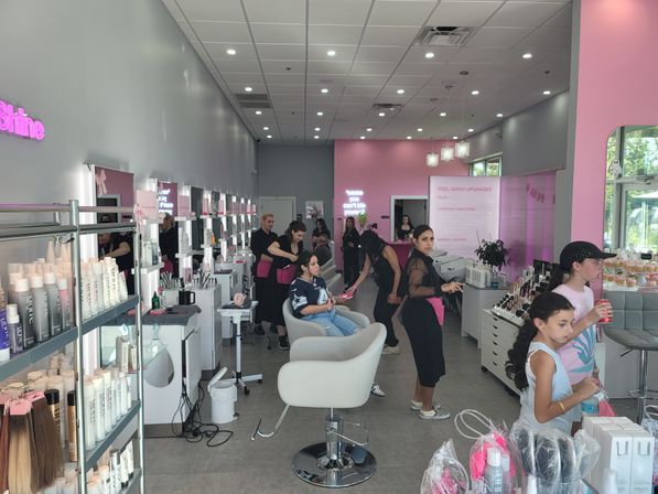 Bright pink-and-gray family hair salon interior with multiple styling stations, stylists working on kids and customers, retail shelves of hair products, and children waiting with drinks.